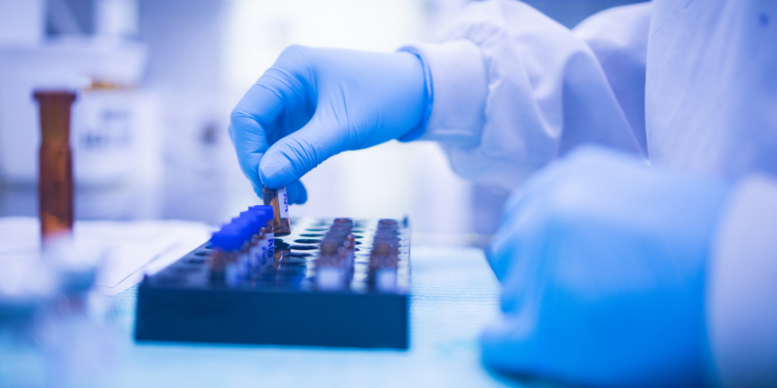 Close-up of a laboratory technician wearing blue gloves and a protective gown, placing small vials into a black tray on a workbench in a clinical or pharmaceutical setting.